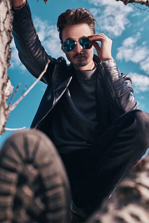 Handsome young man in a black leather jacket and sunglasses sitting in a tree in open air. The spirit of freedom and adventure. Men's fashion.の写真素材