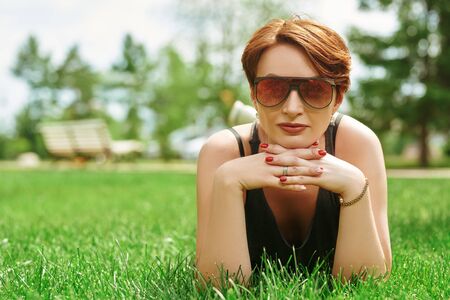 Happy middle-aged woman in modern sunglasses lying relaxing on a green lawn in the summer park. の写真素材