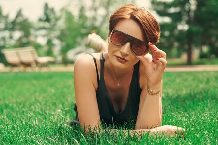 Happy middle-aged woman in modern sunglasses lying relaxing on a green lawn in the summer park. の写真素材