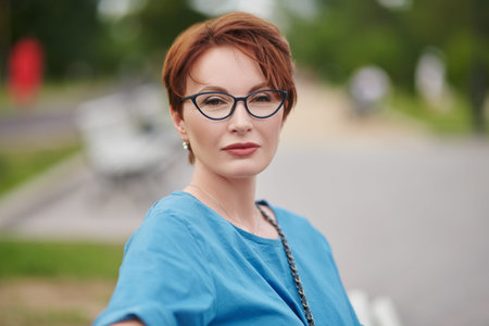 Happy middle-aged woman in elegant glasses and blue dress posing in the summer park. Optics for...