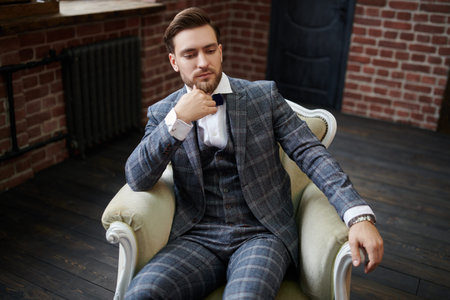 Portrait of a handsome good-looking dreamy man in elegant suit sitting relaxed in an armchair....
