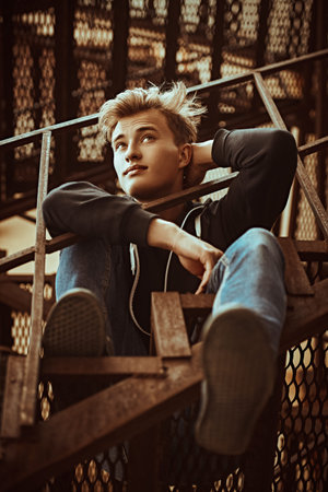 Handsome young man in casual clothes sits on a metal industrial staircase. Urban grunge style. Male...