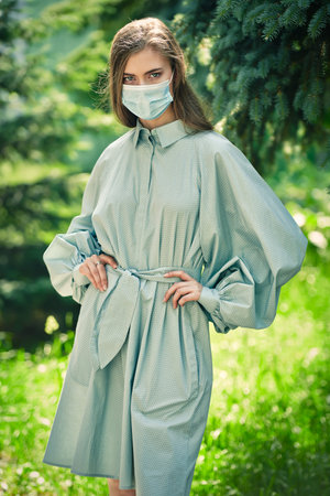 Portrait of a young woman in elegant summer dress and face mask  walking in the park. Sunny summer...