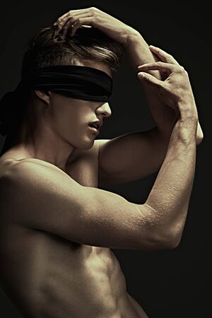 Art portrait of a handsome blindfold young man with perfect muscular body looking up at the light. Black background, darkness.の写真素材