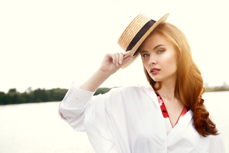 Beautiful romantic girl in a straw hat and a light white shirt stands on the riverbank. Summer holidays.の写真素材