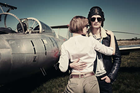 A beautiful girl and a handsome man pilots stand next to a fighter jet at the airfield. Military aircraft. Military fashion. Retro style.の写真素材