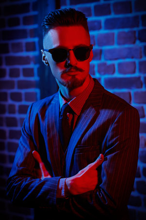 Portrait of a handsome young man in elegant suit and black sunglasses standing in red light by a...