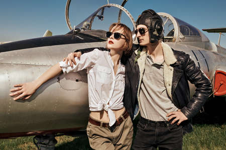 A beautiful girl and a handsome man pilots stand next to a fighter jet at the airfield. Military aircraft. Military fashion. Retro style.の写真素材