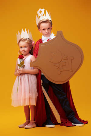Cute boy and girl in costumes of Prince and Princess are posing on on a yellow background. Childhood dreams. Fantasy, imagination. Studio portrait.の写真素材