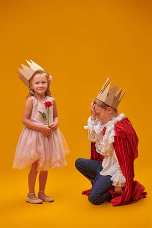 A emotional boy in costume of Prince and gives a rose to a girl in a Princess costume. Childhood dreams. Fantasy, imagination. Studio portrait on a yellow background.の写真素材
