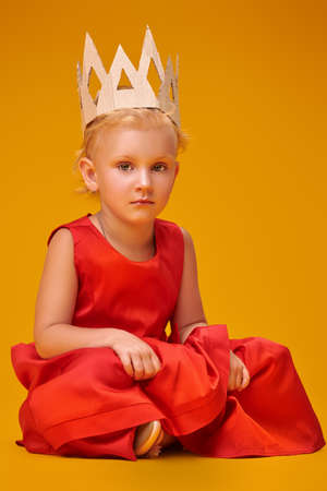 Portrait of a cute little girl posting in red dress and on crown a yellow background. Studio shot. Happy childhood.の写真素材