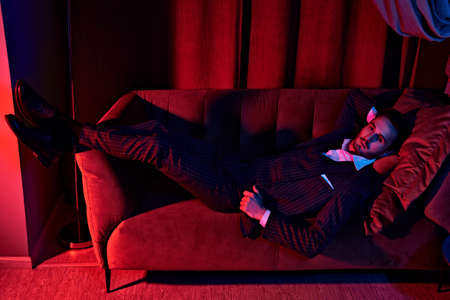 Portrait of a handsome brunet man lying on the sofa in a luxury apartment. Glamorous lifestyle. Fashion shot. Men's clothing and accessories.の写真素材