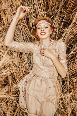 Romantic young woman with beautiful red hair lies on a wheat field and laughs. Beauty, fashion. Modern hippie, bohemian style.の写真素材