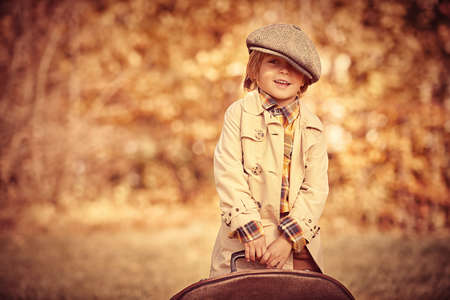 Happy little boy in a coat and cap is carrying a large brown retro suitcase in a beautiful autumn park. Retro style. Children's fashion.の写真素材