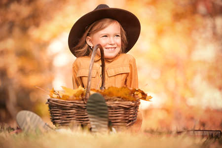 Autumn. Little girl resting, sitting with a basket of autumn leaves on the grass. Retro style. Children's fashion.の写真素材