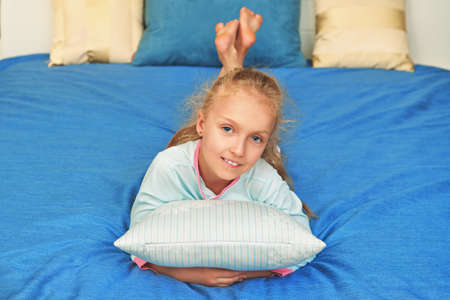 Joyful funny blonde girl lying on a bed hugging a pillow and smiling at camera. Happy childhood. Family at home.の写真素材