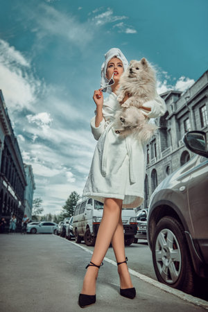 Glamorous lady in a white terry dressing gown with a white towel on her head and high heels walks along the street of a big city with her small fluffy dog. Beauty, fashion.の写真素材
