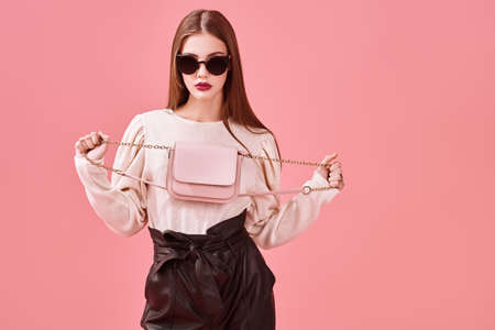 Fashionable young woman posing at studio in modern clothes and sunglasses on a pink background.の写真素材