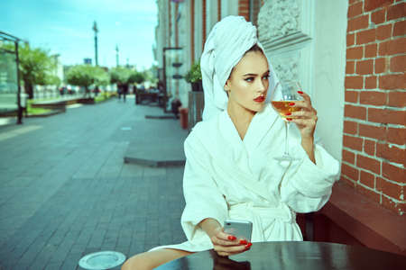 Glamorous young woman in a white terry dressing gown with a white towel on her head sits at a table in a street cafe with a glass of wine and holding a phone in her hand. Fashion shot.の写真素材