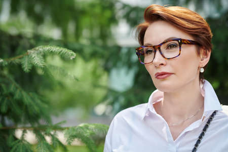 Elegant middle-aged woman in glasses and white shirt stands in the park on a summer day. Business woman portrait. Optics style.の写真素材