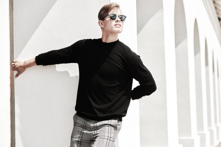 Fashion shot. Handsome young man model in black pullover and black sunglasses posing next to white...