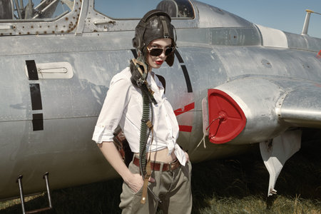 Portrait of a beautiful girl pilot wearing helmet and sunglasses stands next to her fighter jet at...