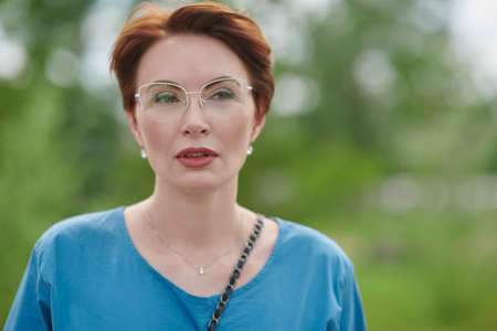 Surprised middle-aged woman in elegant glasses and blue blouse posing in the summer park. Optics for women.の写真素材