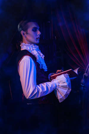 Handsome vampire aristocrat of the 19th century in an elegant suit and with a drop of blood on his lips reading a book in his castle. Halloween.の写真素材