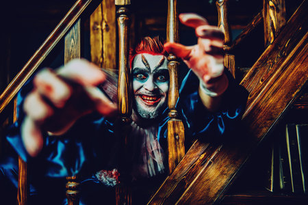 A close up portrait of a smiling clown pulling hands from behind stairs railings. Halloween,...