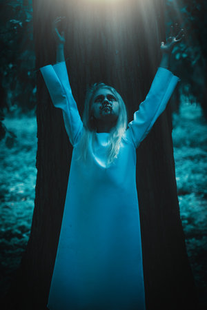 A portrait of a scary pale girl looking up and raising his hands at the light against the background of a tree in the forest. Horror film, Zombie, halloween.の写真素材