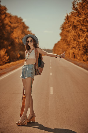 Pretty hippie girl with a guitar and a backpack hitchhiking on a highway. Spirit of freedom. Summer vacation adventure.の写真素材