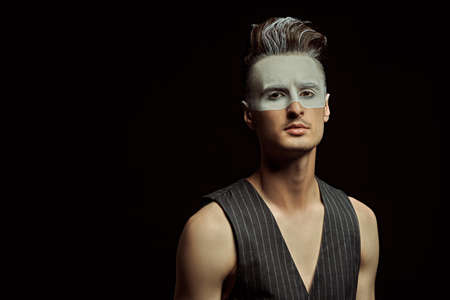 Art portrait of a handsome young man with white paint on his face on a black background with copy space. Male beauty, fashion. Hairstyle and cosmetics.の写真素材