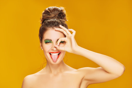 Portrait of an attractive playful girl with colorful makeup posing with expression on a yellow...