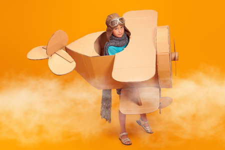 Cute dreamer girl playing with a cardboard airplane. Childhood. Fantasy, imagination. Studio portrait on a yellow background.の写真素材