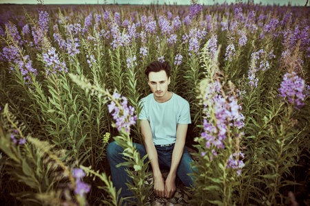 Portrait of a handsome young man in jeans and white t-shirt sitting relaxed in blooming field...