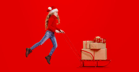 Merry Christmas and Happy New Year! Happy girl child in Christmas hat pushing the sled with gifts....