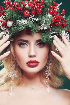 Close-up portrait of a beautiful Christmas woman with a Christmas spruce wreath on her head, decorated with Christmas balls, berries and snowflakes. Winter beauty girl. X-mas makeup and cosmetics.の写真素材