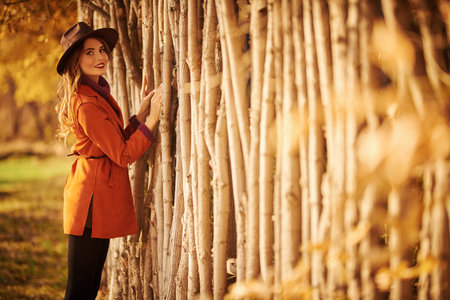 Beautiful young woman stands by a wooden hedge in an autumn park at sunset. Autumn beauty, fashion.