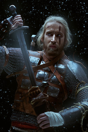 Portrait of a brave medieval warrior in armor with a sword on a snowy background. The Witcher man.