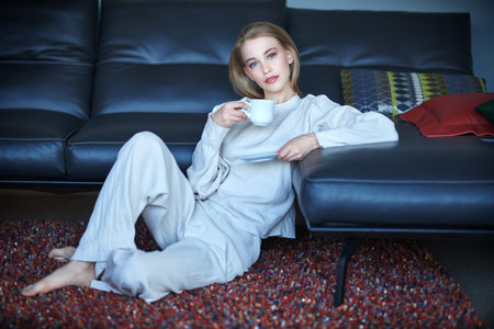 Beautiful happy girl having a rest in her cozy living room, she is sitting on a soft carpet next to the sofa and drinks coffee. Home interior, furniture. Lifestyle.の写真素材