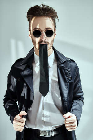 Modern stylish man in elegant black suit, white shirt and sunglasses holding a tie in his mouth on a gray background with shadows. Portrait of a modern businessman. Men's fashion.の写真素材