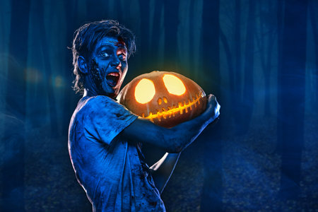 Halloween concept. Portrait of a scary zombie boy teenager holding a pumpkin lantern in forest. Copy space.の写真素材