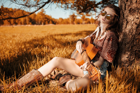 Romance of freedom. Beautiful hippie girl plays the guitar, sitting under a tree in the prairie....