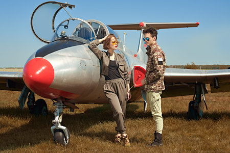 A beautiful girl and a handsome man pilots stand next to a fighter jet at the airfield. Military...