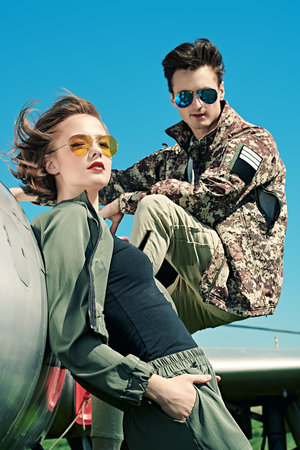 A beautiful girl and a handsome man pilots is posing next to a fighter jet at the airfield. Military...