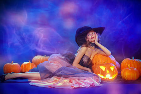 Charming beautiful witch lies with pumpkins lantern in a luxury evening dress and a hat. Halloween celebration, party. Copy space. Halloween fashion.の写真素材