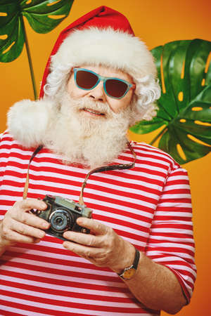 Joyful Santa Claus is relaxing and taking pictures in hot tropical countries. Christmas Holidays, tourist trips to the sea. Bright yellow background.の写真素材