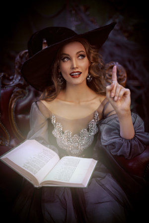 Portrait of a charming woman witch in a hat and beautiful dress sitting in an armchair with a book...