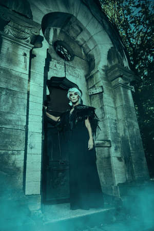 An aristocratic old widow woman with beautiful gray hair and a rich black dress stands sad in a crypt in a cemetery. Black Widow. Atmosphere of mysticism. Halloween.の写真素材