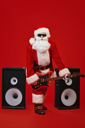 Santa Claus rock musician plays an electric guitar on the background of large speakers. Red festive...